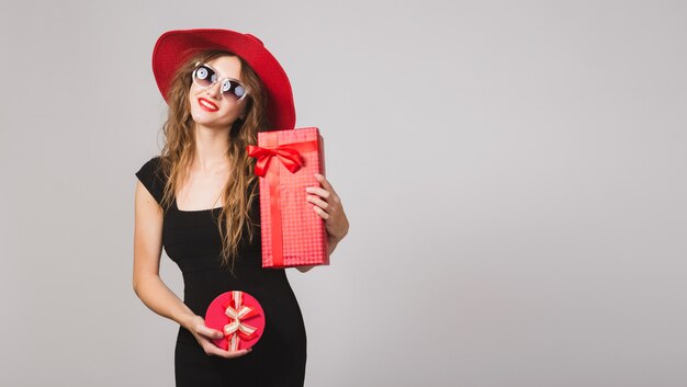 Jovem e bela mulher segurando presentes, vestido preto, chapéu vermelho, óculos de sol, feliz, sorridente, sexy, elegante, caixas de presente, comemorando, positiva