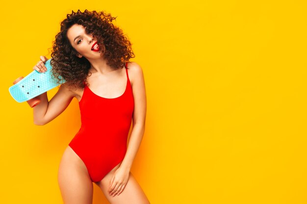 Jovem e bela mulher sorridente posando perto da parede amarela no estúdioModelo sexy em traje de banho vermelhoFêmea positiva com penteado de cachos segurando o skate pennyFeliz e alegre