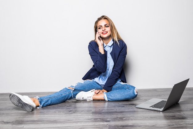 Jovem e bela mulher sorrindo, sentada no chão com o computador falando ao telefone.