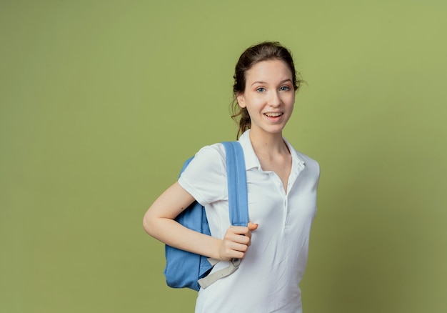 Jovem e bonita aluna alegre vestindo uma bolsa e segurando a alça da bolsa, isolada em um fundo verde oliva com espaço de cópia