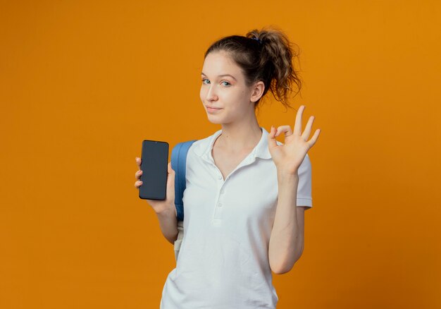 Jovem e bonita aluna confiante usando uma bolsa de costas mostrando o celular e fazendo um sinal de ok isolado em um fundo laranja com espaço de cópia