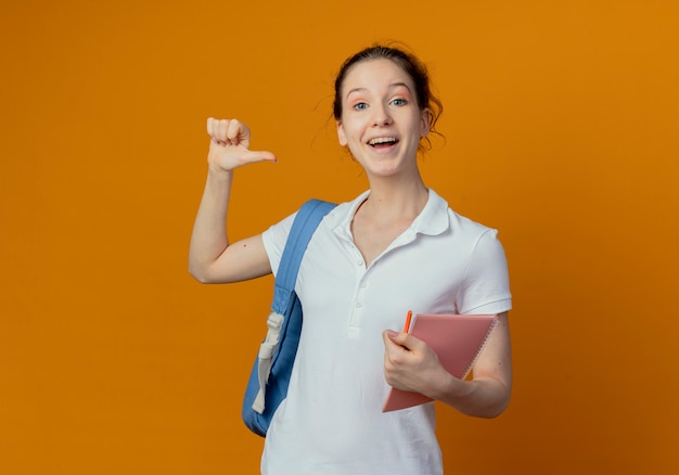 Jovem e bonita aluna impressionada com uma bolsa de costas segurando um bloco de notas e uma caneta apontando para si mesma, isolada em um fundo laranja com espaço de cópia