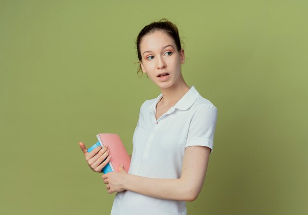 Jovem e bonita aluna impressionada em pé na vista de perfil, segurando um bloco de notas e um livro e olhando para o lado isolado no fundo verde oliva com espaço de cópia