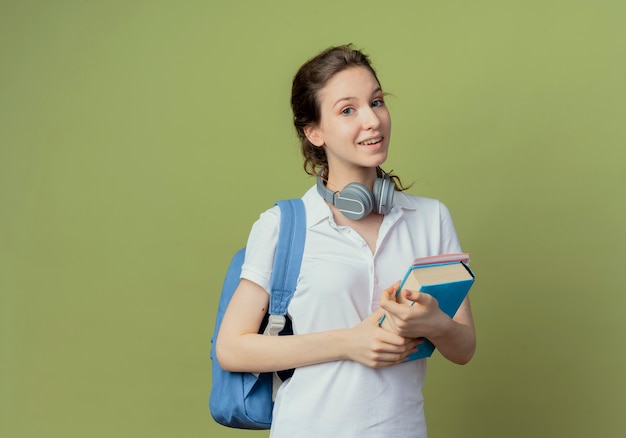 Foto grátis jovem e bonita aluna impressionada usando bolsa nas costas e fones de ouvido no pescoço segurando um livro e um bloco de notas