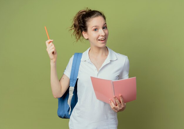 Jovem e bonita aluna impressionada usando uma bolsa de volta segurando um bloco de notas e levantando uma caneta isolada sobre fundo verde oliva