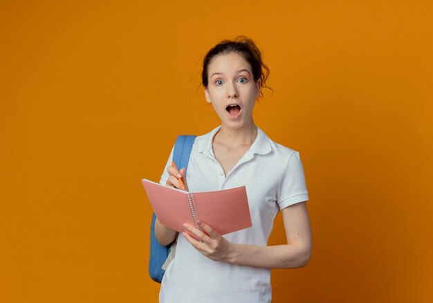 Jovem e bonita aluna impressionada usando uma bolsa de volta segurando uma caneta e um bloco de notas isolado em um fundo laranja com espaço de cópia