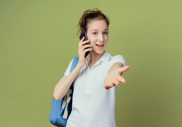 Jovem e bonita aluna impressionada usando uma bolsa traseira, falando ao telefone e estendendo a mão para a câmera, isolada em um fundo verde com espaço de cópia