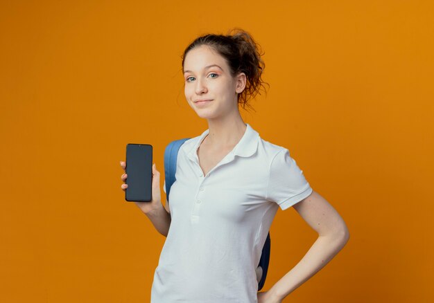 Jovem e bonita aluna satisfeita usando uma bolsa de trás mostrando o celular isolado em um fundo laranja