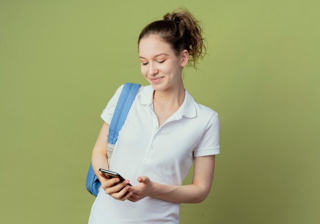 Jovem e bonita aluna satisfeita vestindo uma bolsa de costas, segurando e olhando para o celular, isolado em um fundo verde com espaço de cópia