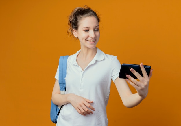 Jovem e bonita aluna sorridente usando uma bolsa de costas segurando e olhando para um telefone celular isolado em um fundo laranja com espaço de cópia