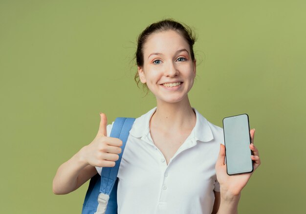 Jovem e bonita aluna sorridente usando uma bolsa de trás mostrando o celular e o polegar isolados no fundo verde