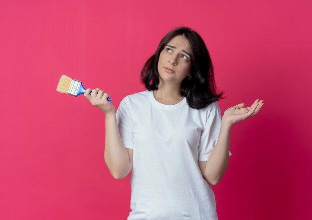 Jovem e bonita caucasiana confusa, olhando para o lado, segurando o pincel e mostrando a mão vazia, isolada no fundo carmesim