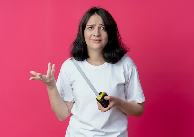 Foto grátis jovem e bonita caucasiana confusa segurando o medidor de fita e mostrando a mão vazia, isolada em um fundo carmesim com espaço de cópia