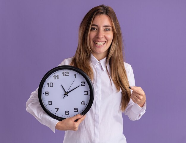 Jovem e bonita caucasiana sorridente segura e aponta para o relógio roxo