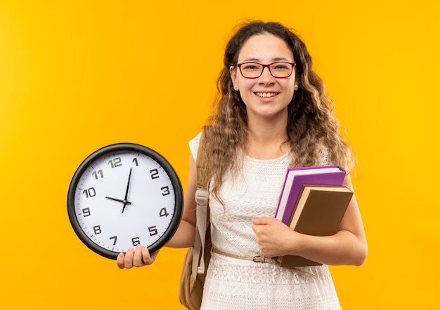Foto grátis jovem e bonita estudante sorridente usando óculos e uma bolsa traseira segurando um relógio e livros isolados na parede amarela