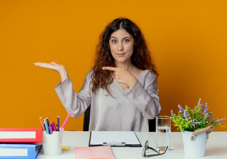 Foto grátis jovem e bonita funcionária de escritório satisfeita sentada à mesa com ferramentas de escritório, fingindo estar segurando algo isolado em laranja