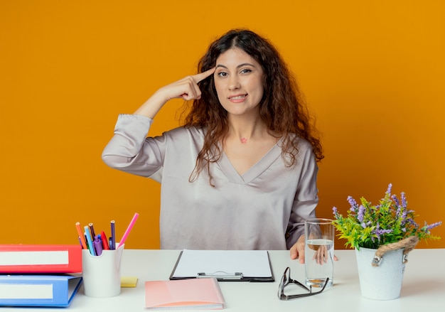 Jovem e bonita trabalhadora de escritório impressionada, sentada à mesa com ferramentas de escritório, colocando o dedo na testa, isolada na parede laranja