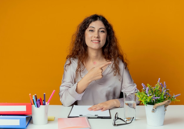 Jovem e bonita trabalhadora de escritório sorridente, sentada à mesa com as ferramentas de escritório apontando para o lado isolado em laranja com espaço de cópia