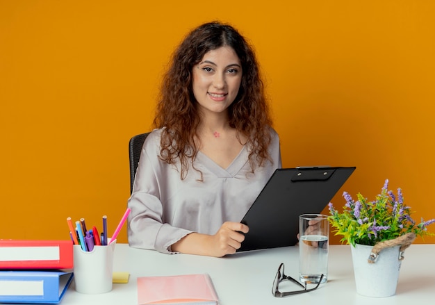 Jovem e bonita trabalhadora de escritório sorridente, sentada à mesa com ferramentas de escritório, segurando a prancheta