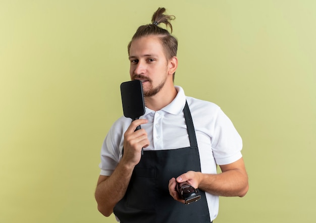 Jovem e bonito barbeiro satisfeito segurando o pente e a tesoura de cabelo fingindo cantar usando o pente como microfone isolado na parede verde oliva