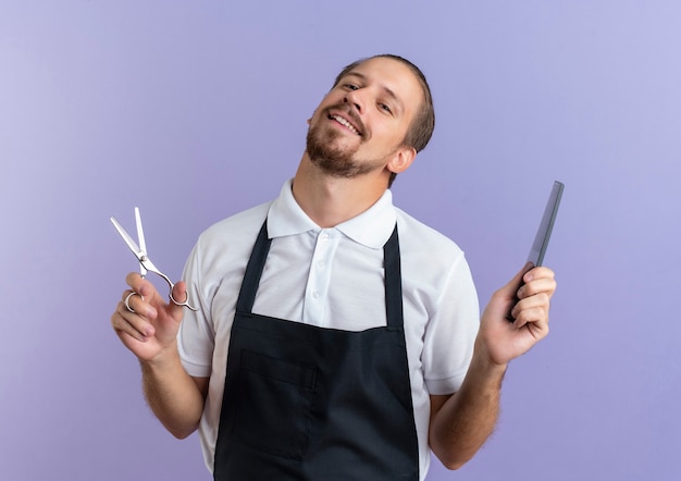 Jovem e bonito barbeiro sorridente, usando uniforme, segurando uma tesoura e um pente isolado na parede roxa