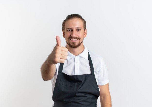 Jovem e bonito barbeiro sorridente, vestindo uniforme, mostrando o polegar isolado na parede branca