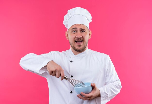 Jovem e bonito cozinheiro impressionado em uniforme de chef segurando um batedor e uma tigela isolados na parede rosa