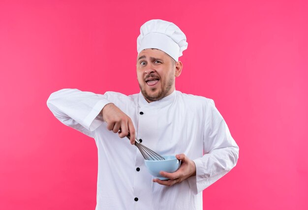 Jovem e bonito cozinheiro impressionado em uniforme de chef segurando uma tigela e um batedor isolado na parede rosa