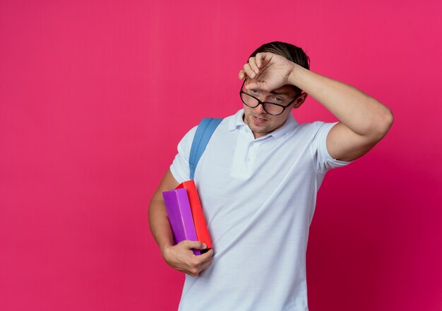 Jovem e bonito estudante cansado com uma bolsa nas costas e óculos segurando livros