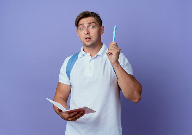 Jovem e bonito estudante do sexo masculino impressionado usando uma bolsa de costas segurando um caderno e levantando uma caneta isolada na parede azul