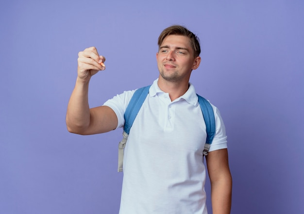 Jovem e bonito estudante do sexo masculino satisfeito usando a bolsa, segurando e olhando para uma caneta isolada no azul
