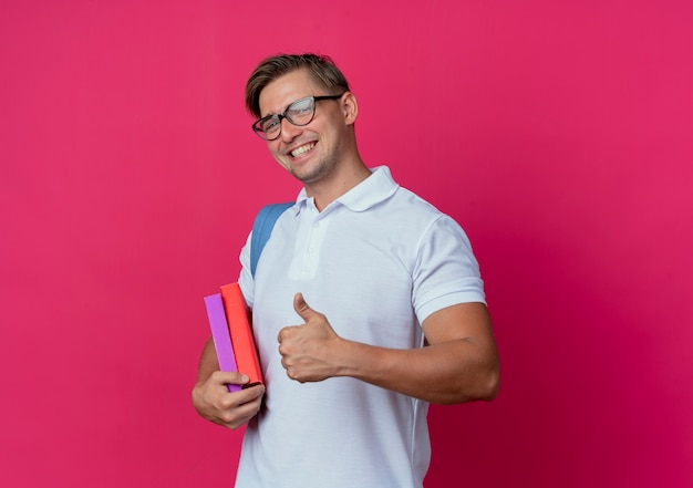 Foto grátis jovem e bonito estudante do sexo masculino sorridente usando bolsa e óculos segurando livros