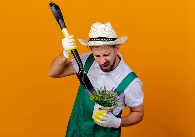 Foto grátis jovem e bonito jardineiro eslavo de uniforme, irritado, usando chapéu e luvas de jardinagem, segurando uma pá e um vaso de flores espadeando flores em um vaso isolado