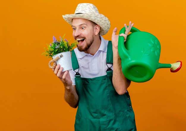 Jovem e bonito jardineiro eslavo impressionado de uniforme e chapéu segurando um vaso de flores e um regador olhando para um vaso de flores isolado na parede laranja com espaço de cópia