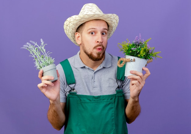 Foto grátis jovem e bonito jardineiro eslavo impressionado de uniforme e chapéu segurando vasos de flores olhando