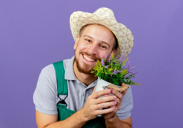 Jovem e bonito jardineiro eslavo sorridente de uniforme e chapéu segurando um vaso de flores, parecendo isolado na parede roxa com espaço de cópia