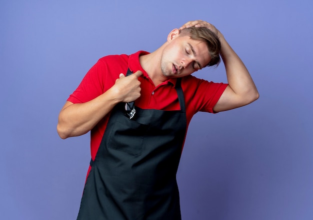 jovem e cansado barbeiro loiro com os olhos fechados vestindo uniforme coloca a mão na cabeça segurando uma máquina de cortar cabelo