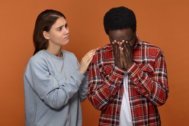 Foto grátis jovem e compassiva linda esposa branca confortando e consolando o infeliz marido negro que está chorando por causa de sérios problemas no trabalho. mulher europeia carinhosa apoiando o namorado africano