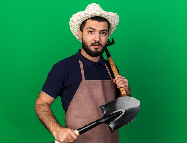 Jovem e confiante jardineiro caucasiano com chapéu de jardinagem, segurando o ancinho e a pá isolados na parede verde com espaço de cópia