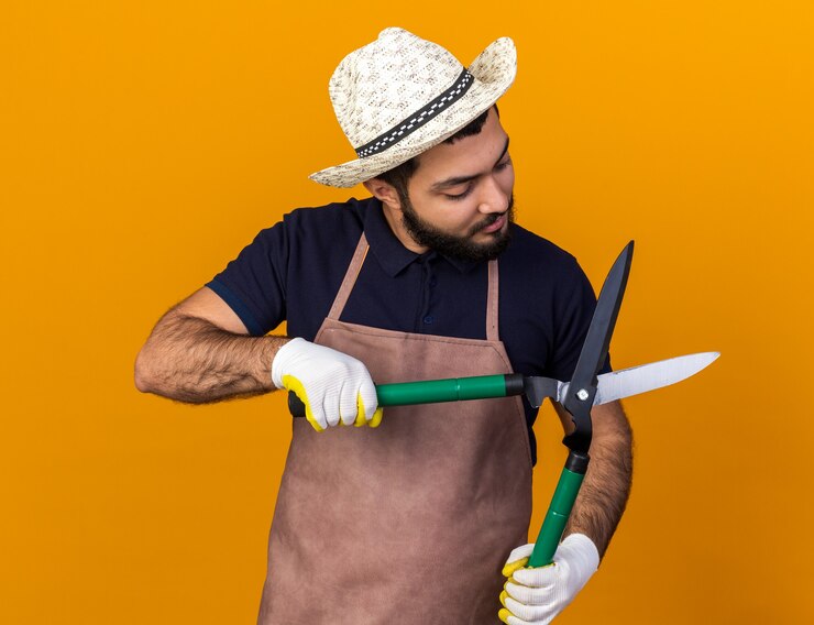 Foto grátis jovem e confiante jardineiro caucasiano com chapéu e luvas de jardinagem, segurando e olhando para uma tesoura de jardinagem isolada na parede laranja com espaço de cópia