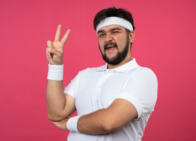 Jovem e desportivo piscando usando bandana e pulseira, mostrando um gesto de paz isolado na parede rosa