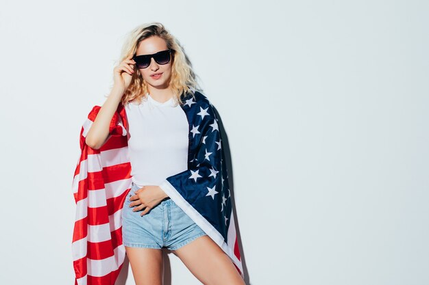 Jovem e despreocupado. Mulher jovem e bonita loira carregando a bandeira americana e sorrindo enquanto celebra o dia 4 de julho, o Dia da Independência em pé contra uma parede branca