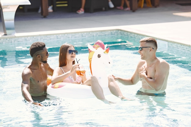 Jovem e dois amigos multirraciais do sexo masculino relaxando em uma piscina. Menina vestindo maiô branco e óculos escuros