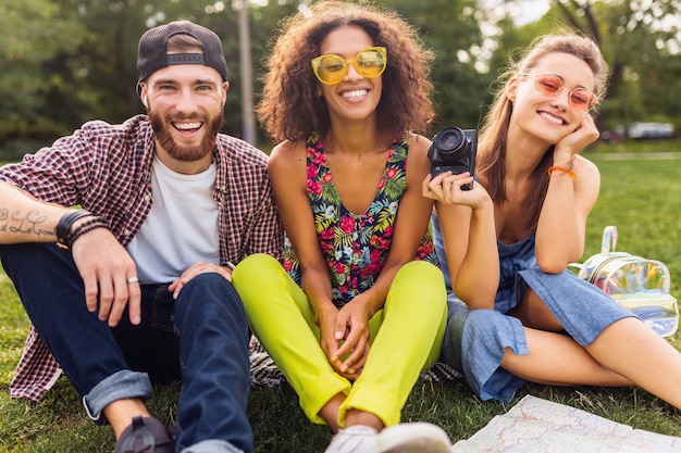 Jovem e feliz companhia de amigos sorridentes sentados no parque, homens e mulheres se divertindo juntos, viajando com a câmera
