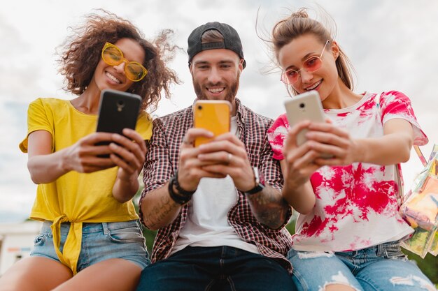 Jovem e feliz companhia de amigos sorridentes sentados no parque usando smartphones