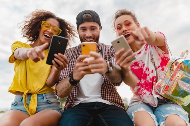 Jovem e feliz companhia de amigos sorridentes sentados no parque usando smartphones