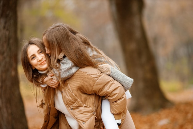Jovem e garotinha na floresta de outono Mulher carrega sua filha nas costas Garota usando vestido cinza fashion com uma jaqueta