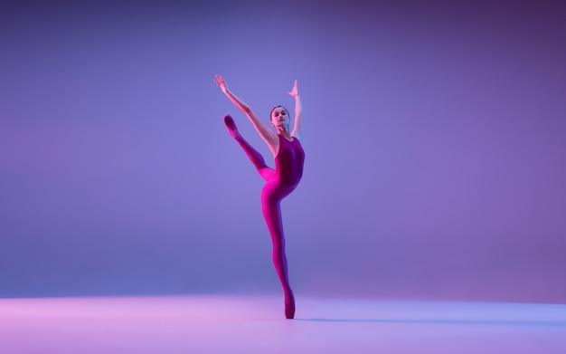 Jovem e graciosa dançarina de balé isolada no fundo roxo do estúdio em luz de néon