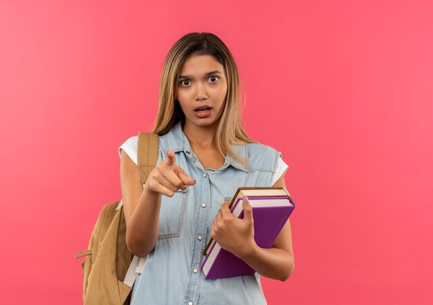 Jovem e linda aluna impressionada com uma bolsa nas costas, segurando livros e apontando para a frente, isolada em rosa