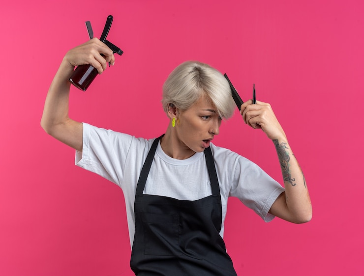 Foto grátis jovem e linda barbeira feminina de uniforme segurando ferramentas de barbeiro, penteando o cabelo isolado na parede rosa